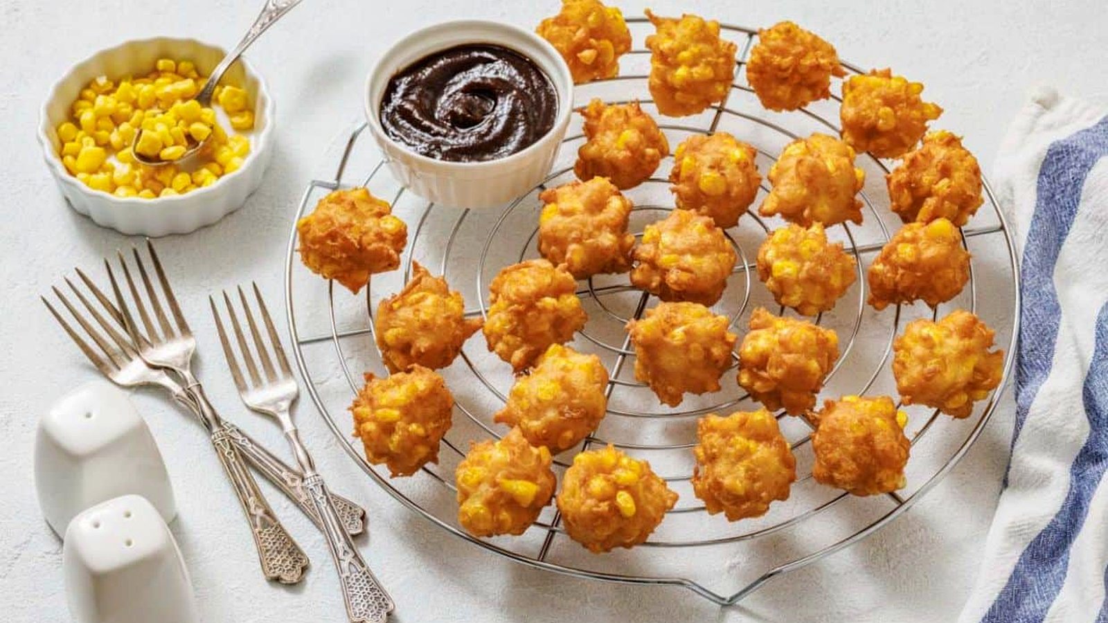 A cooling rack with corn nuggets, a bowl of dipping sauce, a bowl of corn, three forks, a spoon, and salt and pepper shakers on a light surface.