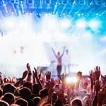 A crowd of people with their hands raised at a neon city festival, facing a brightly lit stage.