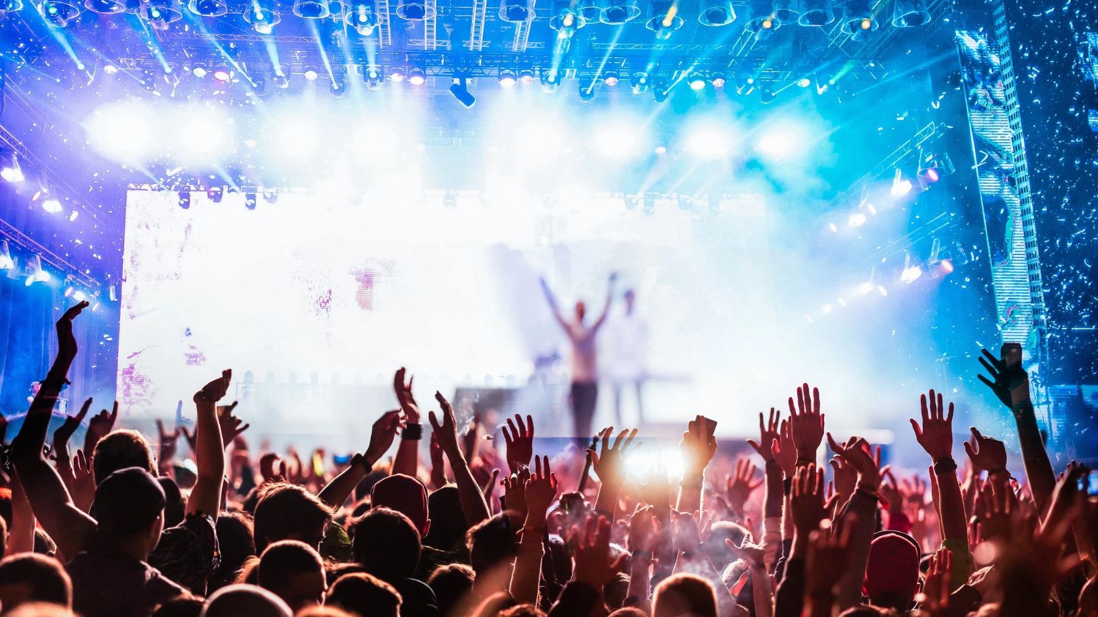 A crowd of people with their hands raised at a neon city festival, facing a brightly lit stage.
