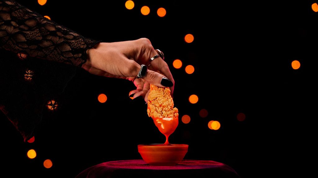 A hand with black-painted fingernails dips a breaded chicken tender into a small bowl of orange sauce against a dark background with orange bokeh lights.