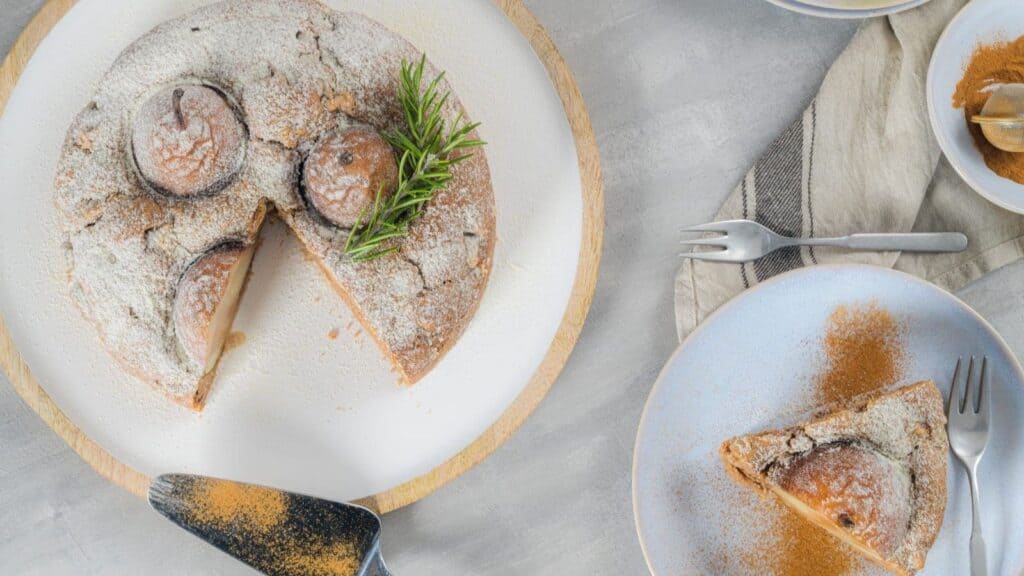 A pear cake dusted with powdered sugar on a wooden board, served as a fall dessert recipe without pumpkin, with a single slice on a plate and a small bowl of cinnamon nearby.