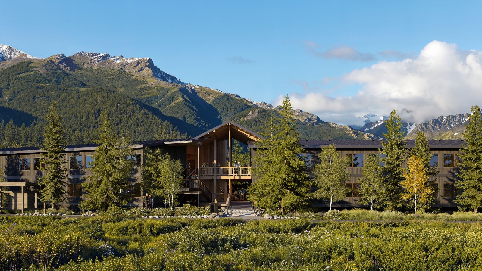 A modern two-story hotel building with a central gabled porch, set in front of mountains with snow-capped peaks.