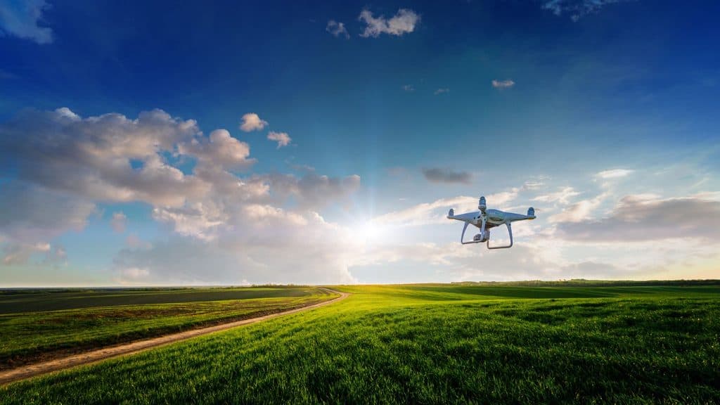 A white quadcopter drone flying over a green field with a dirt path under a bright sky.