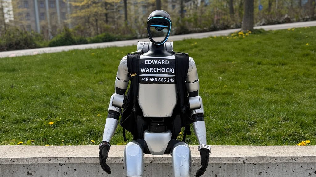 A silver humanoid robot sitting on a concrete ledge, wearing a black vest with &ldquo;EDWARD WARCHOCKI&rdquo; and a phone number printed on the chest.