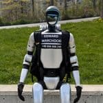 A silver humanoid robot sitting on a concrete ledge, wearing a black vest with “EDWARD WARCHOCKI” and a phone number printed on the chest.