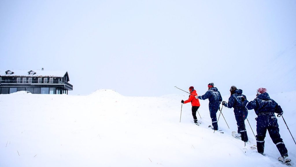 Four people in winter gear cross a snowy slope toward a house in the distance.