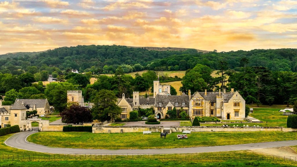 Large stone country manor house with a circular driveway and manicured lawns set against a backdrop of rolling hills and forest.