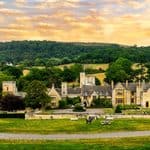 Large stone country manor house with a circular driveway and manicured lawns set against a backdrop of rolling hills and forest.