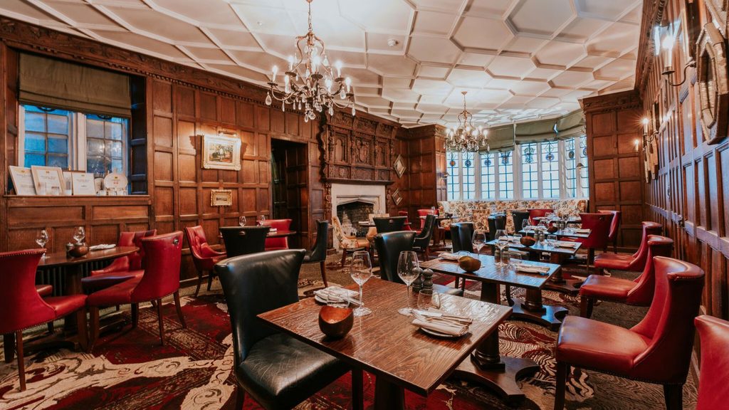 Dining room with wood-paneled walls, crystal chandeliers, a coffered ceiling, and tables set with leather chairs and red upholstery.