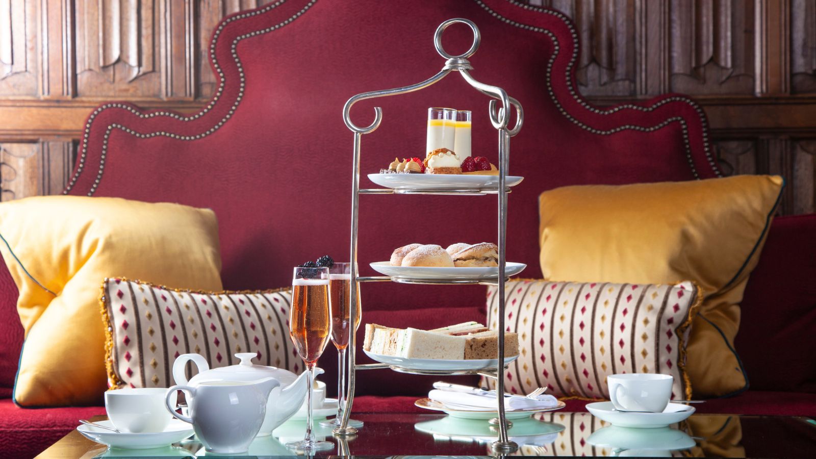 A three-tiered afternoon tea stand served with finger sandwiches, scones, and small desserts, accompanied by glasses of sparkling wine and a teapot.