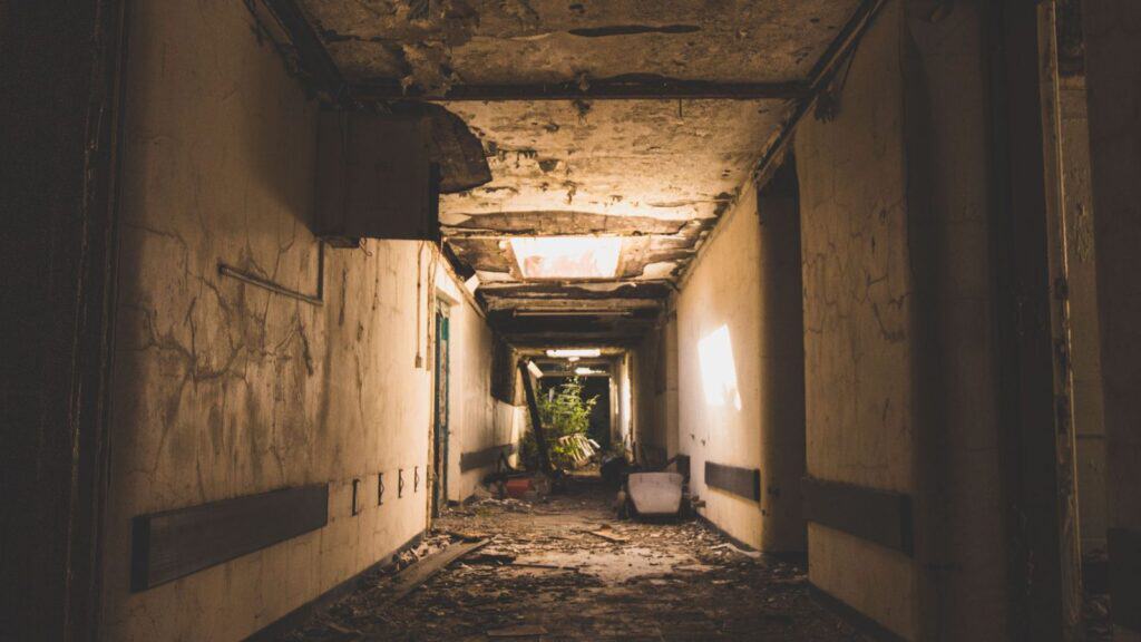 A dilapidated, debris-filled hallway in a decaying building, perfect for a spookiest small-town fall road trip, with peeling paint and sunlight streaming through holes in the ceiling.
