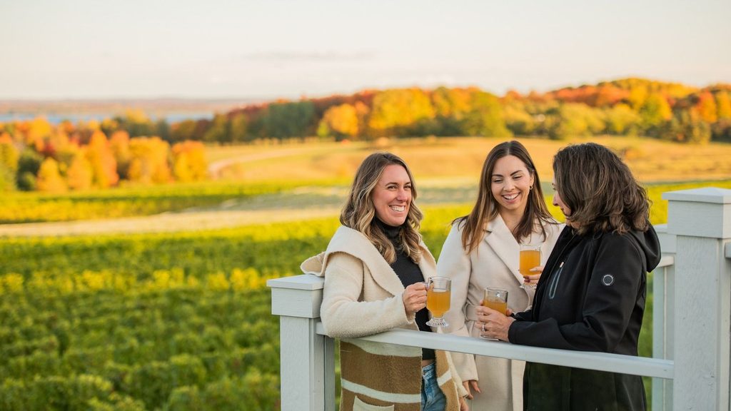 Three women holding glasses of cider on a deck overlooking a vineyard during autumn.