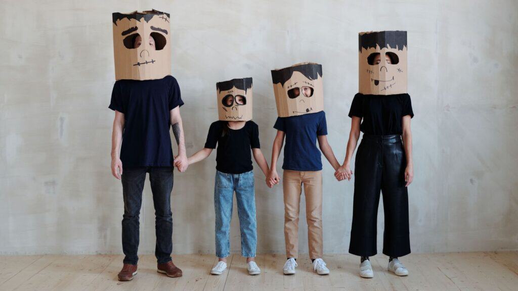 Four people standing in a row in family Halloween costumes at home, holding hands and wearing cardboard box masks decorated like Frankenstein’s monster.