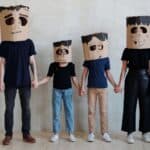 Four people standing in a row in family Halloween costumes at home, holding hands and wearing cardboard box masks decorated like Frankenstein’s monster.