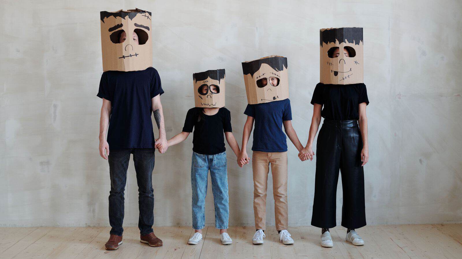 Four people standing in a row in family Halloween costumes at home, holding hands and wearing cardboard box masks decorated like Frankenstein’s monster.