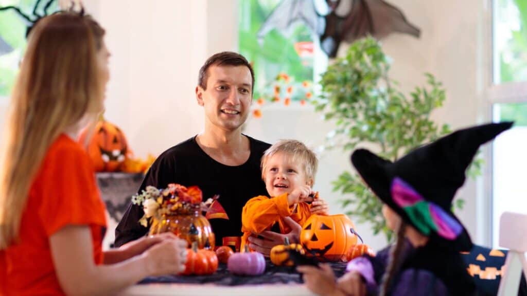 A family in Halloween costumes sitting at a table decorated with pumpkins as a toddler plays with tricks.