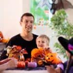 A family in Halloween costumes sitting at a table decorated with pumpkins as a toddler plays with tricks.