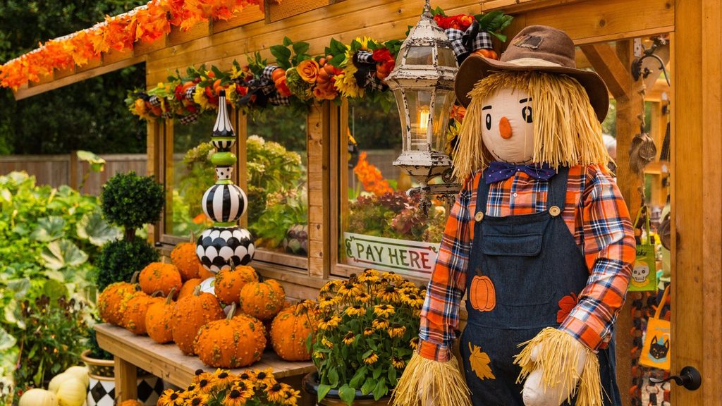 A scarecrow stands by a fall farm stand, displaying pumpkins, autumn garlands, and a sign that reads &ldquo;Pay Here.&rdquo;
