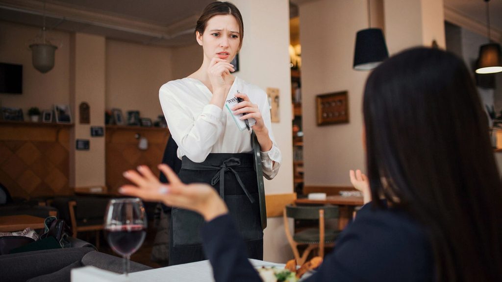 A server holding a notepad listens with a concerned expression as a customer, perhaps celebrating National Grouch Day, speaks to her across a restaurant table.