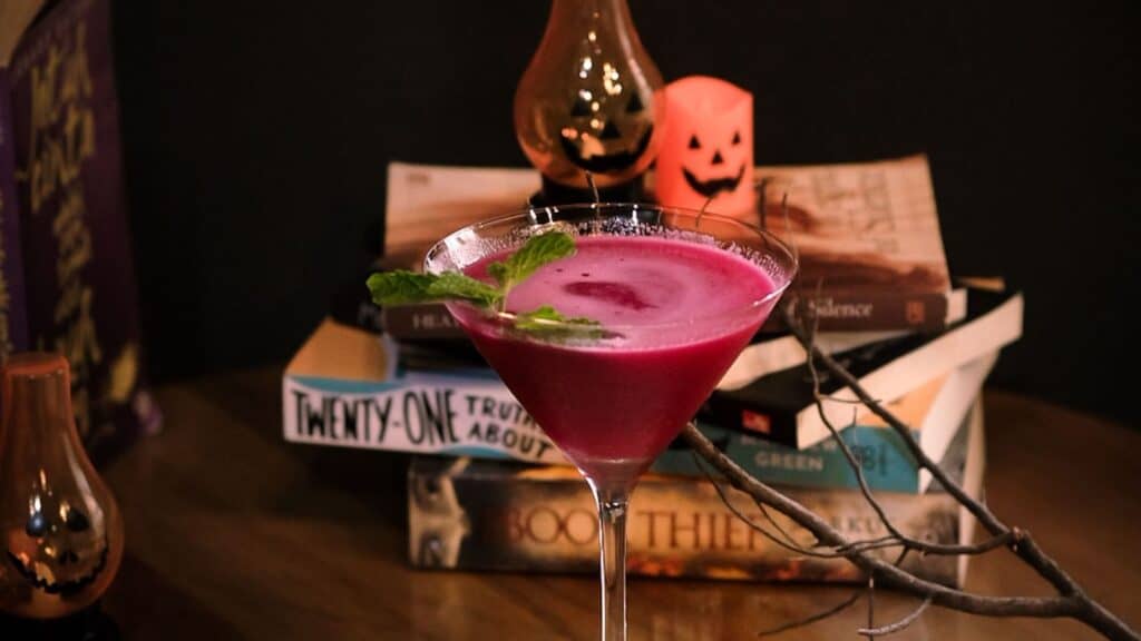 A pink Halloween cocktail in a martini glass garnished with mint, set in front of a stack of books and festive decorations.