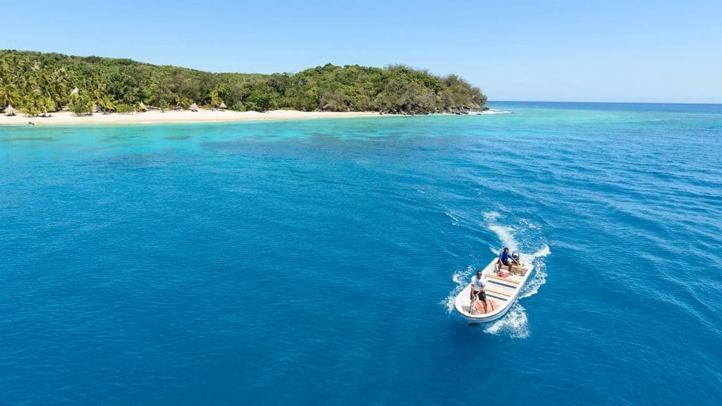 A small motorboat with two people traveling across clear blue water toward a tropical beach.