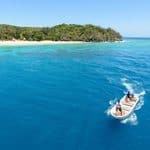 A small motorboat with two people traveling across clear blue water toward a tropical beach.