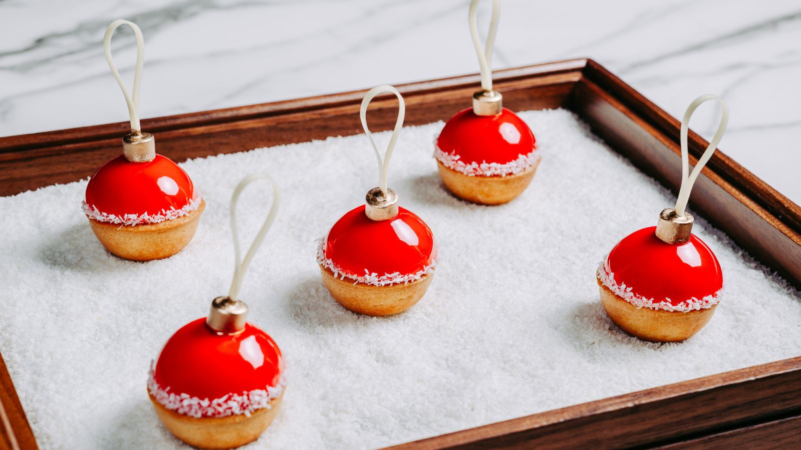 Five small tarts topped with red, spherical ornaments on a wooden tray filled with shredded coconut, reminiscent of holiday Las Vegas classes.