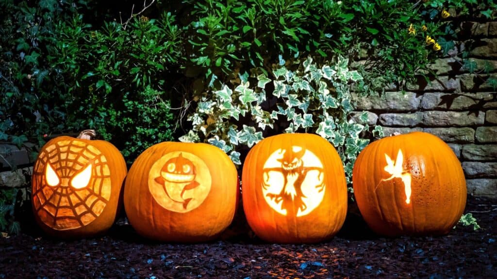 Four pumpkins carved with advanced pumpkin carving techniques display designs of Spider-Man, a shark, a spooky figure, and Tinker Bell lined up in front of a hedge.