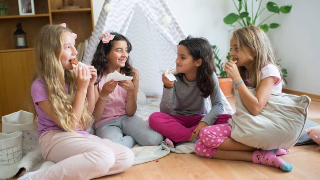 Four children in family Halloween costumes sitting on the floor in a playroom, eating pizza.