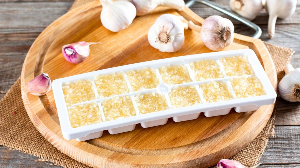 Minced garlic frozen in a white ice cube tray on a wooden cutting board surrounded by whole garlic bulbs.