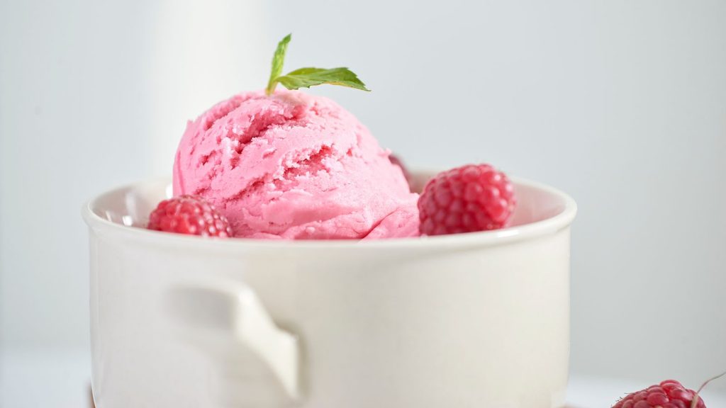 A scoop of pink raspberry ice cream topped with a mint leaf and fresh raspberries in a white bowl.