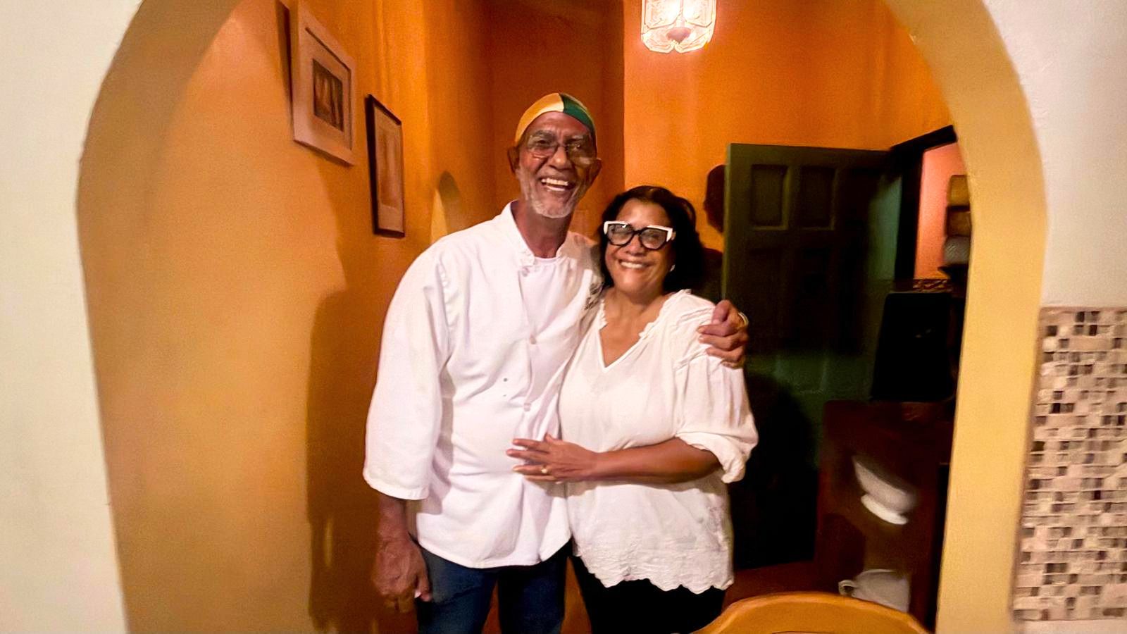 A smiling man in a chef’s coat and a woman stand together in a doorway with their arms around each other, capturing a moment of Grenada food travel.