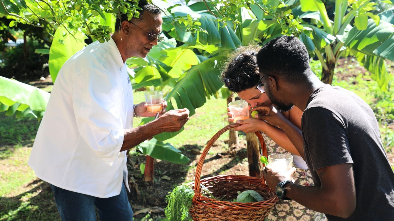 A chef in a white coat shows two people how to taste ingredients held in a wicker basket during a Grenada food travel experience in a garden.