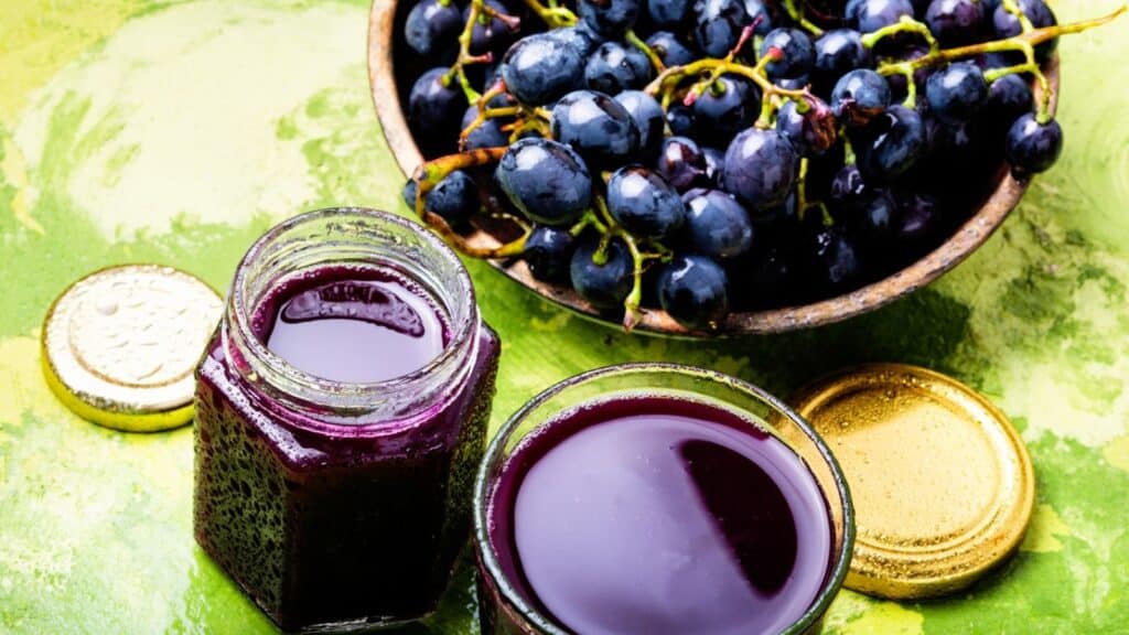 A bowl of fresh black grapes next to a jar and a glass filled with dark purple grape juice, representing fall harvest foods worth preserving.