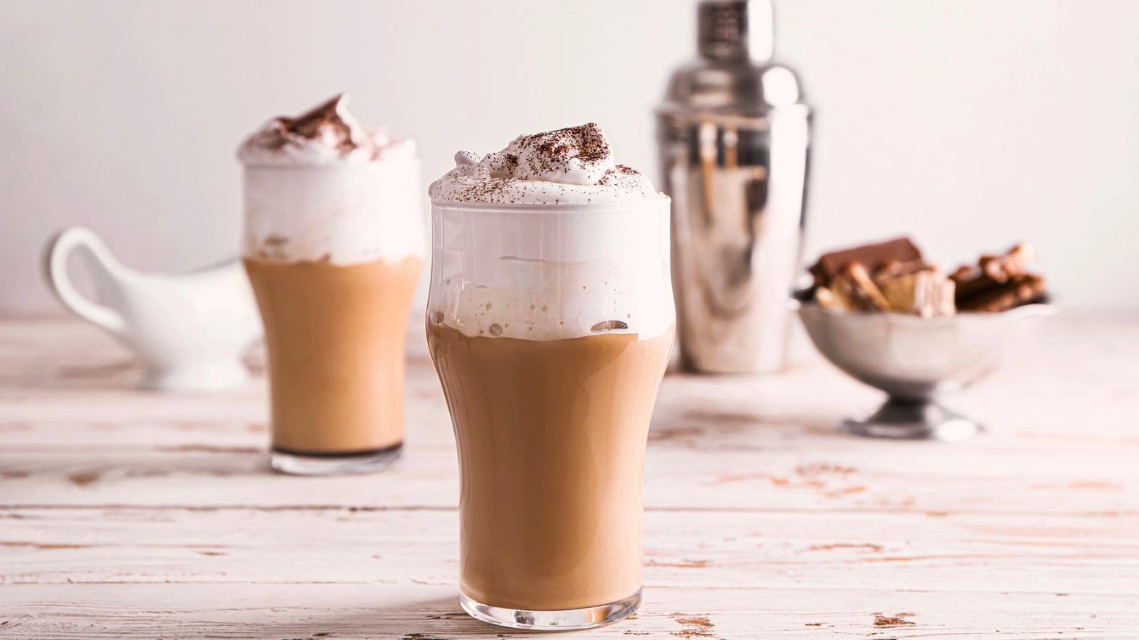 Two glasses of iced coffee topped with whipped cream and cocoa powder, with a cocktail shaker and a bowl of chocolate pieces in the background.