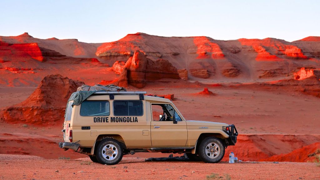 A tan off-road vehicle with &ldquo;DRIVE MONGOLIA&rdquo; printed on the side, parked in a red, rocky desert landscape.