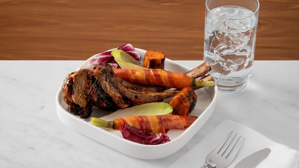 A plate of grilled lamb chops with bacon-wrapped carrots and radicchio, next to a glass of water and cutlery.