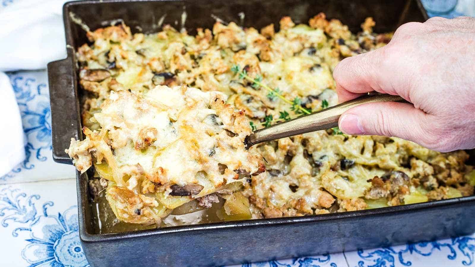 A hand lifts a serving of French Country Ground Pork and Potato Casserole from a rectangular metal pan on a patterned surface.