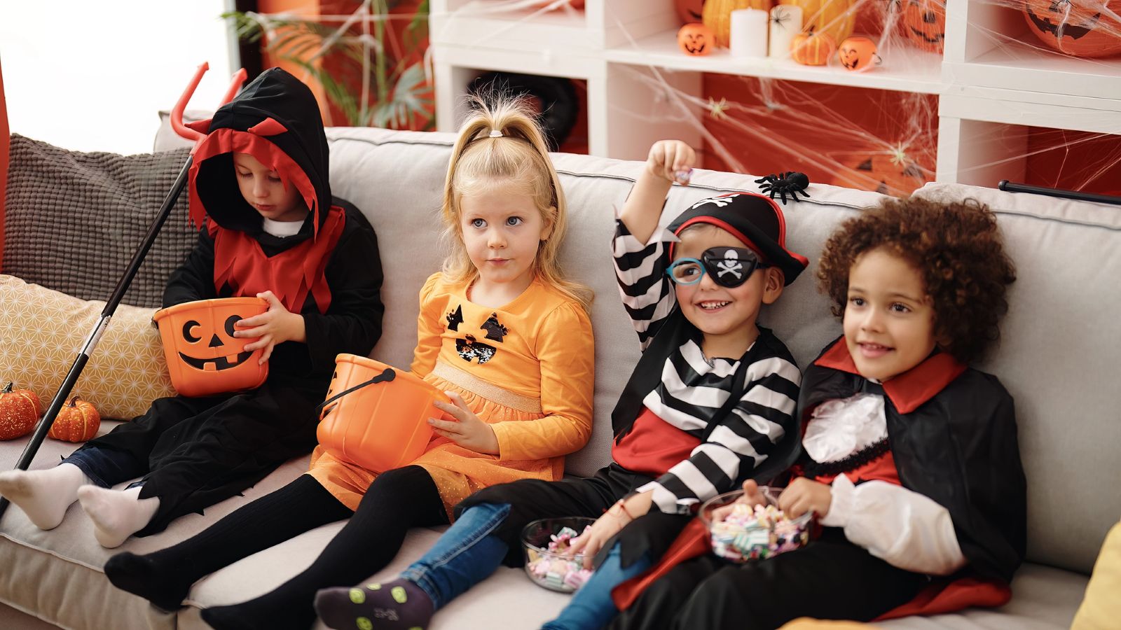 Four children in Halloween costumes sit on a couch holding plastic jack-o’-lantern buckets and bowls of candy while learning how to make Halloween candy last longer.