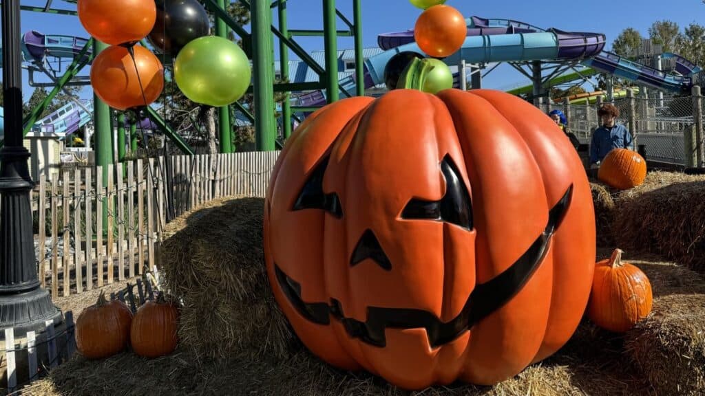 A large jack-o&rsquo;-lantern decoration sits on hay bales in front of a water slide at a theme park with Halloween events, surrounded by pumpkins and orange, black, and green balloons.
