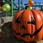 A large jack-o’-lantern decoration sits on hay bales in front of a water slide at a theme park with Halloween events, surrounded by pumpkins and orange, black, and green balloons.
