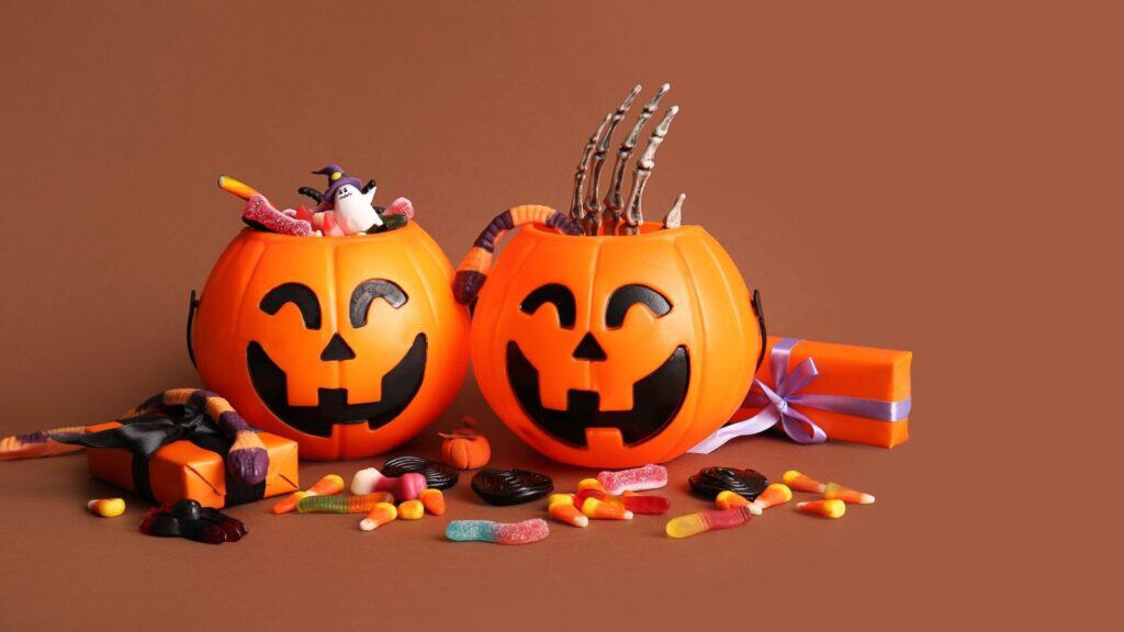 Two orange plastic jack-o&rsquo;-lantern candy bowls filled with assorted sweets and a skeleton hand prop, surrounded by scattered Halloween tricks and two wrapped gift boxes.