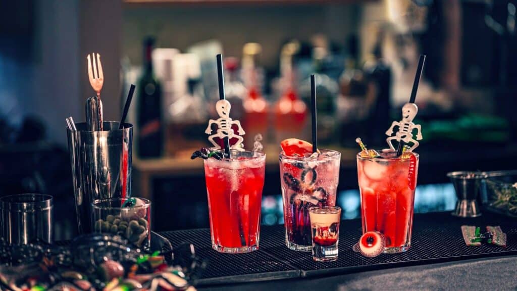 Three Halloween cocktails served in glasses on a bar counter, each decorated with skeleton swizzle sticks and festive garnishes.