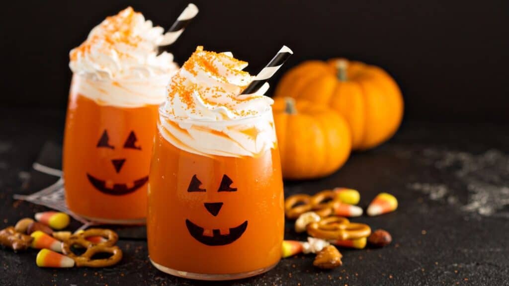 Two glasses of orange Halloween punch decorated with jack-o&rsquo;-lantern faces, topped with whipped cream and sprinkles, served as candy corn treats alongside pretzels.