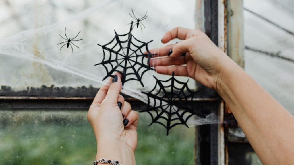 Hands placing paper spiderwebs and small spider cutouts made from the recycling bin onto a window covered in decorative faux cobwebs.