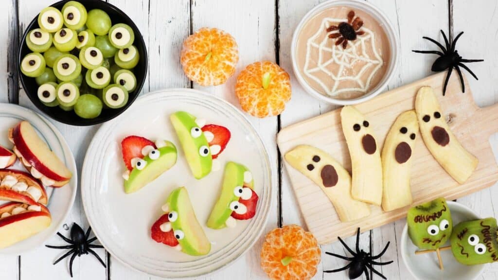 Halloween-themed trunk-or-treat snack ideas include green grapes with candy eyes, apple slices with strawberry mouths, banana ghosts, tangerine pumpkins, and a bowl of dip decorated with a spiderweb.