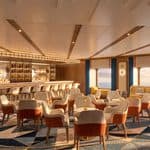 A cruise ship lounge with a lit bar, high-top stools, and clusters of chairs and small tables on a patterned rug, facing windows with an ocean view.
