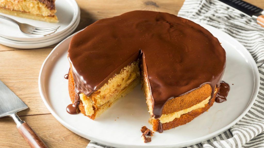 A yellow sponge cake with custard filling and a chocolate ganache topping, pictured for National Boston Cream Pie Day with one slice removed.