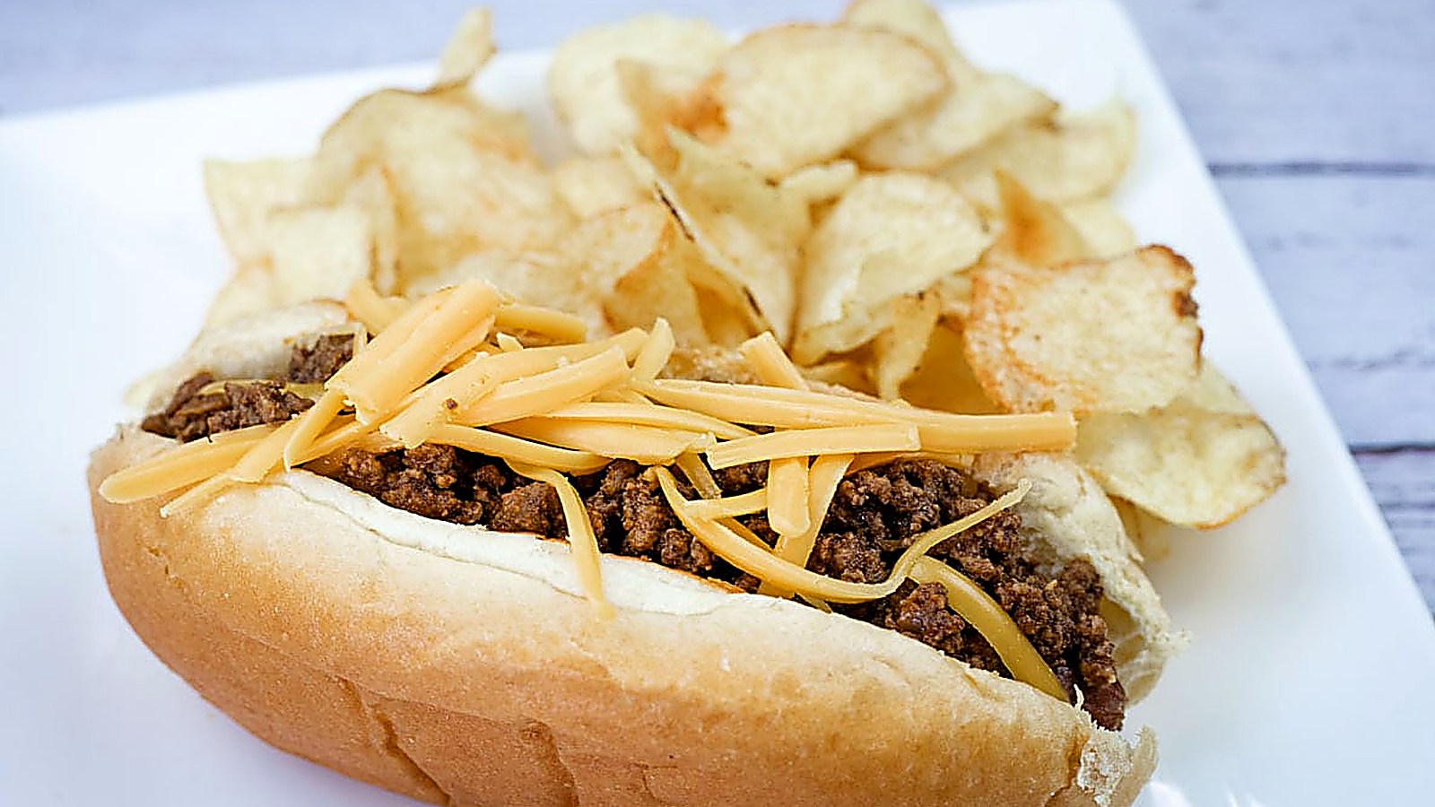 A hot dog chili topped with shredded cheddar, served with potato chips on a white plate.