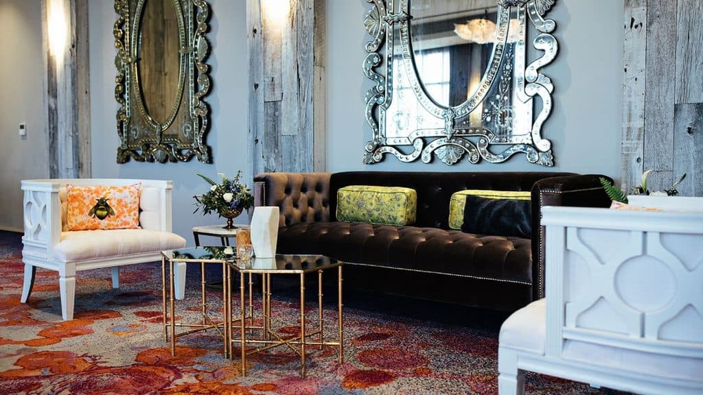A dark brown tufted sofa with yellow cushions, two white accent chairs, and two mirrored coffee tables in a room with ornate mirrors and a patterned rug.